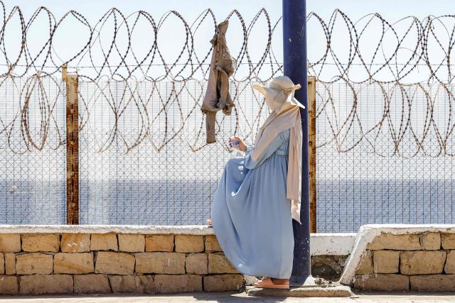 A woman sits by a barbed-wire fence as relatives and friends wait for the return of people who had previously crossed into Spain's North African enclave of Ceuta, at Morocca's Bab Sebta border crossing on the outskirts of Fnideq on August 2, 2026. As many as 60,000 migrants entered Ceuta in recent days, mostly swimming around the small border post that juts into the Mediterranean Sea, in an unprecedented surge that prompted an international crisis for Spain. The mass crossing into the tiny Spanish territory resulted in at least 67 deaths, according to Spanish authorities, and led to clashes with Moroccan police on the shared border. (Photo by Abdel Majid BZIOUAT / AFP)