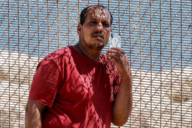 A man holds a phone while standing next to a fence as relatives and friends wait for the return of people who had previously crossed into Spain's North African enclave of Ceuta, at Morocca's Bab Sebta border crossing on the outskirts of Fnideq on August 2, 2026. As many as 60,000 migrants entered Ceuta in recent days, mostly swimming around the small border post that juts into the Mediterranean Sea, in an unprecedented surge that prompted an international crisis for Spain. The mass crossing into the tiny Spanish territory resulted in at least 67 deaths, according to Spanish authorities, and led to clashes with Moroccan police on the shared border. (Photo by Abdel Majid BZIOUAT / AFP)