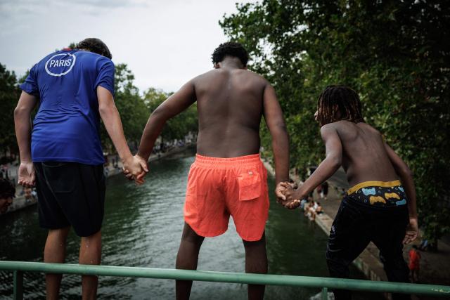 Youths hold hands as they prepare to dive into the Canal Saint-Martin in Paris on August 3, 2026. Authorities have stepped up penalties for people swimming outside designated bathing areas or entering the water when a red flag is flying, under a decree published in France's Official Journal that came into force on August 2, 2026. The measure applies nationwide and is intended to strengthen enforcement of coastal and inland water safety rules amid the peak summer holiday season. (Photo by Simon WOHLFAHRT / AFP)