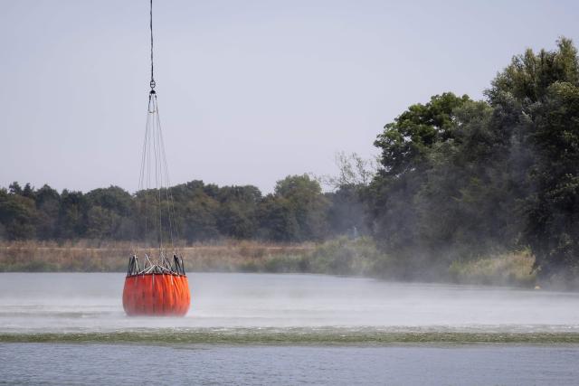 Bystanders watch as a Bambi Bucket suspended beneath a Royal Netherlands Air Force CH-47 Chinook helicopter collects water from the River Meuse in Geijsteren, to battle a wildfire sweeping through the Boschhuizerbergen nature reserve near Oostrum, in the Dutch municipality of Venray, south-eastern Netherlands on August 4, 2026. (Photo by Sem van der Wal / ANP / AFP) / Netherlands OUT