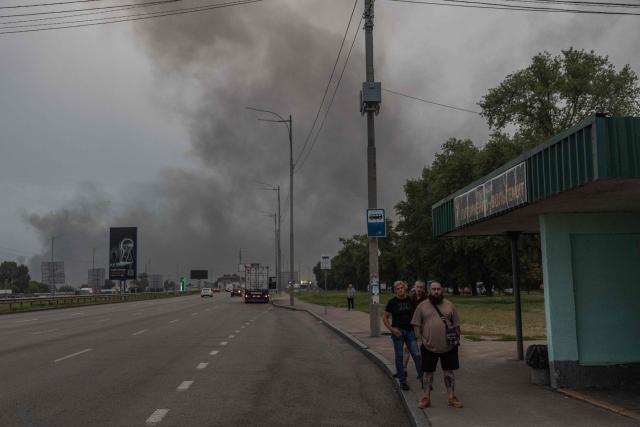 People wait at a bus stop as smoke rising in the distance following massive Russian ballistic missile and drone strikes in Kyiv on August 5, 2026, amid the Russian invasion of Ukraine. Russian strikes on Ukraine's capital and surrounding region killed at least two people and wounded dozens more, authorities said on August 5, 2026, after fresh overnight missile and drone attacks. (Photo by Roman PILIPEY / AFP)