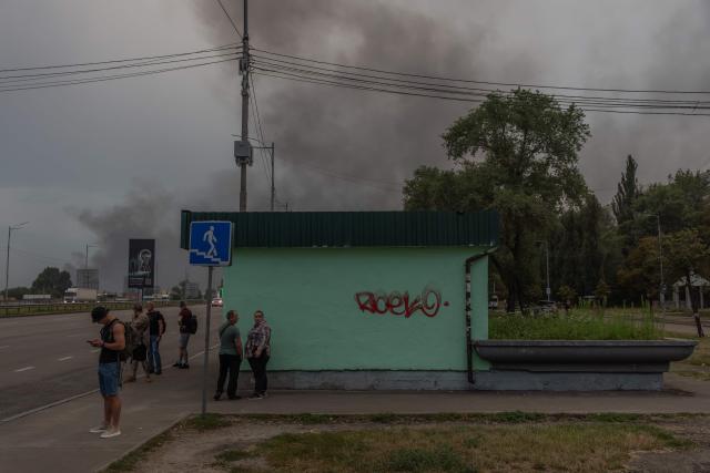People wait at a bus stop as smoke rising in the distance following massive Russian ballistic missile and drone strikes in Kyiv on August 5, 2026, amid the Russian invasion of Ukraine. Russian strikes on Ukraine's capital and surrounding region killed at least two people and wounded dozens more, authorities said on August 5, 2026, after fresh overnight missile and drone attacks. (Photo by Roman PILIPEY / AFP)