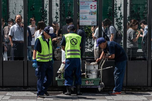 Municipal workers, wearing do not smoke on their vests, clean up trash next to a smoking enclosure in Tokyo on August 6, 2026. (Photo by Andrew CABALLERO-REYNOLDS / AFP)