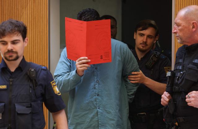 Afghan defendant identified as Farhad N (C), accused of deliberately steering a car into a 1,400-strong trade union march in February 2025, covers his face with a folder as he is escorted by judicial officers into the courtroom as the verdict is expected in his trial, at the Higher Regional Court in Munich, southern Germany on August 6, 2026. The  German court on August 6 handed a life jail term to the Afghan man for a car-ramming attack in Munich last year which killed a two-year-old girl and her mother and left dozens injured. The Munich court ruled that the crime carried "particular gravity", a designation that will make it much harder for the 25-year-old man, named only as Farhad N., to ever win release from prison. (Photo by Karl-Josef HILDENBRAND / AFP)