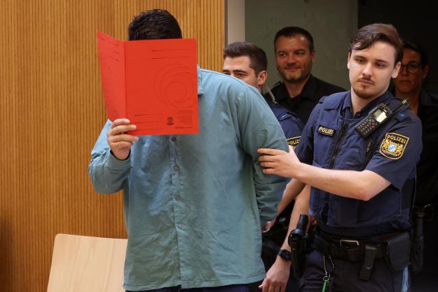 Afghan defendant identified as Farhad N (L), accused of deliberately steering a car into a 1,400-strong trade union march in February 2025, covers his face with a folder as he is escorted by judicial officers into the courtroom as the verdict is expected in his trial, at the Higher Regional Court in Munich, southern Germany on August 6, 2026. The  German court on August 6 handed a life jail term to the Afghan man for a car-ramming attack in Munich last year which killed a two-year-old girl and her mother and left dozens injured. The Munich court ruled that the crime carried "particular gravity", a designation that will make it much harder for the 25-year-old man, named only as Farhad N., to ever win release from prison. (Photo by Karl-Josef HILDENBRAND / AFP)