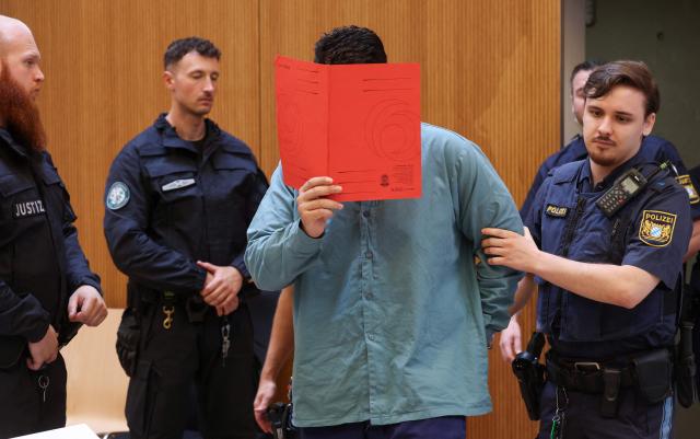 Afghan defendant identified as Farhad N (C), accused of deliberately steering a car into a 1,400-strong trade union march in February 2025, covers his face with a folder as he is escorted by judicial officers into the courtroom as the verdict is expected in his trial, at the Higher Regional Court in Munich, southern Germany on August 6, 2026. A verdict is due on August 6 for the Afghan man accused of a car-ramming attack in Munich last year which killed a two-year-old girl and her mother and left dozens injured. He was 24 at the time. Prosecutors have demanded he be given a life sentence with added conditions to make early release much less likely. The attack was one of several committed by foreign nationals in the run-up to last year's general election that inflamed debate on immigration and security. (Photo by Karl-Josef HILDENBRAND / AFP)