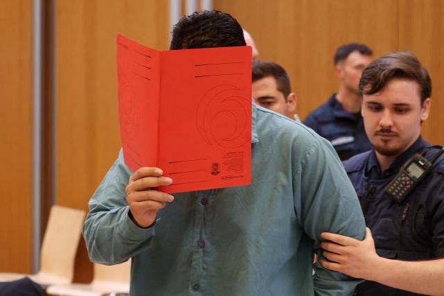 Afghan defendant identified as Farhad N, accused of deliberately steering a car into a 1,400-strong trade union march in February 2025, covers his face with a folder as he is escorted by judicial officers into the courtroom as the verdict is expected in his trial, at the Higher Regional Court in Munich, southern Germany on August 6, 2026. The German court on August 6 handed a life jail term to the Afghan man for a car-ramming attack in Munich last year which killed a two-year-old girl and her mother and left dozens injured. The Munich court ruled that the crime carried "particular gravity", a designation that will make it much harder for the 25-year-old man, named only as Farhad N., to ever win release from prison. (Photo by Karl-Josef HILDENBRAND / AFP)