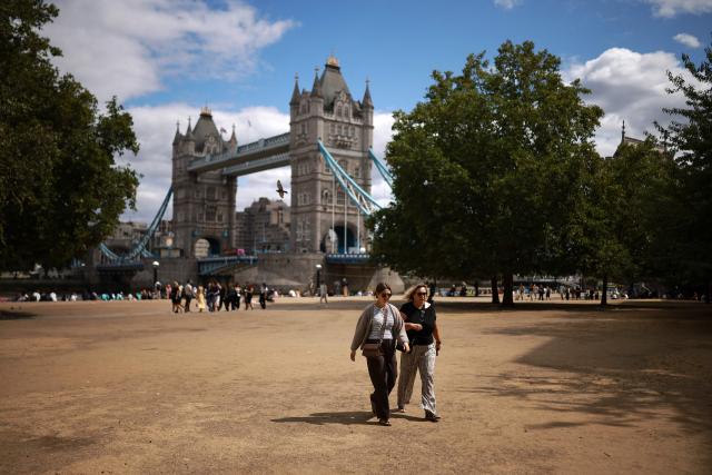 People walk across dry parched grass, turned yellow-brown due to a lack of rain, near Tower Bridge in London on August 6, 2026. London and its suburbs are one of 19 English regions to have had no more than one millimetre of rainfall in July, with water companies in parts of England and Wales imposing bans on using hose pipes for household tasks and watering plants. (Photo by Henry NICHOLLS / AFP)