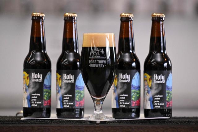 A photo taken on July 2, 2026 shows Robe Town Brewery's beer called Holy Smokes, where brewer Maris Biezaitis feeds leftover grain residue to camels on the Humpalicious camel farm before collecting their dung to be used as fuel to smoke malted barley in the brewing process of the beer, in the South Australian town of Robe. Far from their origins in the Arabian desert, hundreds of thousands of camels roam outback Australia where they are used for wild races, an alternative milk source, and even to make beer. (Photo by William WEST / AFP) / To go with AFP story Australia-camels-animal, by  Xiao Yu Yin