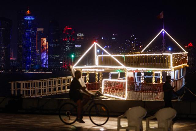 A man rides a bicycle past an illuminated boat along the Corniche Promenade facing the skyline of illuminated high-rise buildings in Doha on August 8, 2026. (Photo by KARIM JAAFAR / AFP)