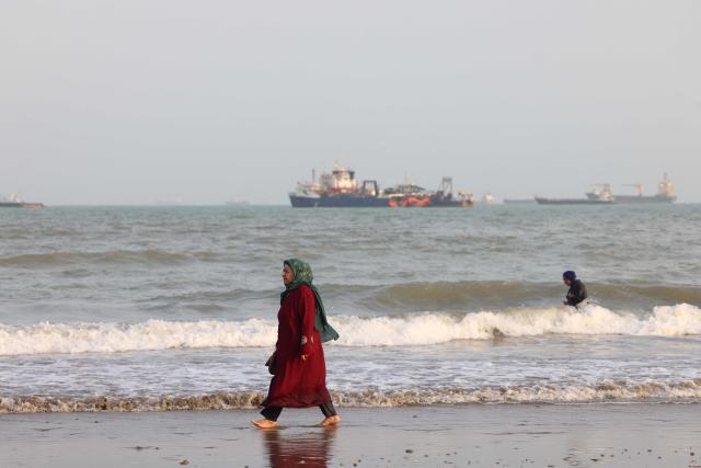 An Iranian woman walks along the seashore as vessels are seen in the Strait of Hormuz off Bandar Abbas, in southern Iran on August 10, 2026. The US President signalled on August 9 that he was prepared to let economic pressure mount against Iran, apparently backing away from more military strikes after Tehran issued a list of demands for opening the Strait of Hormuz. His remarks came after Iran issued over the weekend a list of conditions to reopen the Strait of Hormuz, which has remained largely closed since the start of the Middle East war in late February. (Photo by Atta KENARE / AFP) / 