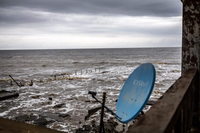 A satellite dish is seen overlooking buildings destroyed by coastal erosion from the King's Palace in Ayetoro, Ondo State, on August 6, 2026. The coastal community of Ayetoro, in Nigeria's southwestern Ondo State, has become one of the country's most visible examples of the vulnerability of communities living along the Atlantic Ocean to coastal erosion and rising waters. Since the early 2000s, coastal erosion in Ayetoro has progressively intensified, with the Atlantic Ocean gradually encroaching on the settlement, swallowing homes, schools, and other infrastructure, and forcing some residents to relocate several times. The ocean that the community has lived alongside for generations has become a constant threat, leaving families to rebuild further inland only to see the water reach them again. For many in Ayetoro, the question is no longer whether the sea will reach their home, but when. (Photo by OLYMPIA DE MAISMONT / AFP)