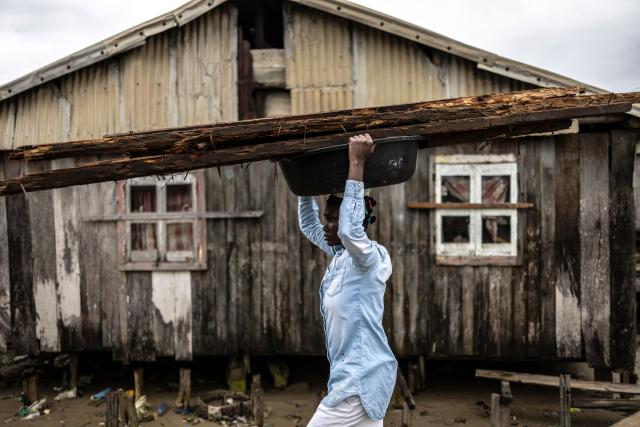 A resident carries wooden planks on a plastic basin on her head past a house affected by coastal erosion in Ayetoro, Ondo State, on August 6, 2026. The coastal community of Ayetoro, in Nigeria's southwestern Ondo State, has become one of the country's most visible examples of the vulnerability of communities living along the Atlantic Ocean to coastal erosion and rising waters. Since the early 2000s, coastal erosion in Ayetoro has progressively intensified, with the Atlantic Ocean gradually encroaching on the settlement, swallowing homes, schools, and other infrastructure, and forcing some residents to relocate several times. The ocean that the community has lived alongside for generations has become a constant threat, leaving families to rebuild further inland only to see the water reach them again. For many in Ayetoro, the question is no longer whether the sea will reach their home, but when. (Photo by OLYMPIA DE MAISMONT / AFP)