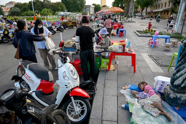 A girl rests on the ground as vendors sell food at Wat Botum park in Phnom Penh on August 11, 2026.  (Photo by TANG CHHIN Sothy / AFP)