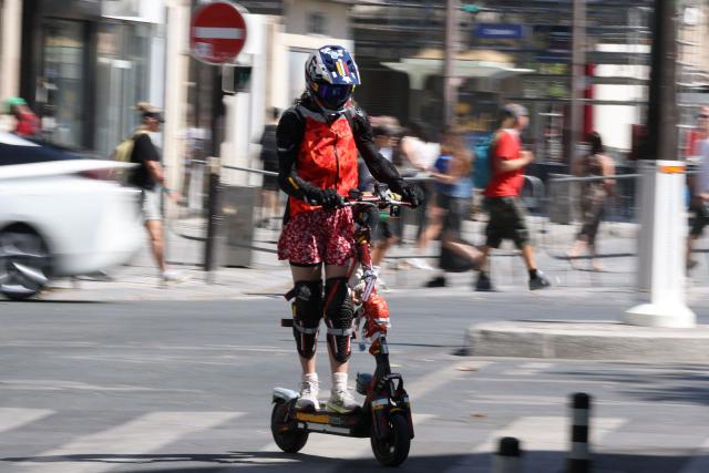 A woman with a helmet rides an electric scooter past traffic on a street of Paris on August 11, 2026. Electric scooter riders in Paris must now wear helmets and reflective gear under new rules introduced after a significant rise in injuries involving the two-wheelers, police said on August 8, 2026. (Photo by Kenzo TRIBOUILLARD / AFP)