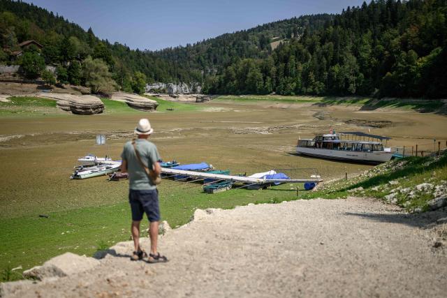 A tourist faces he parched bed of Brenets Lake (Lac des Brenets), part of the Doubs River, a natural border between eastern France and western Switzerland, as central Europe is hit by a new heatwave, in Les Brenets on August 11, 2026. The river has dried up due to a combination of factors, including geological fissures that drain water underground, below-average rainfall and repeated heatwaves. (Photo by Fabrice COFFRINI / AFP)