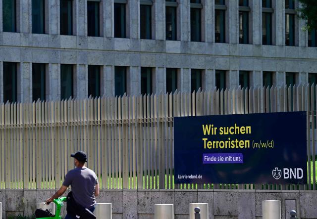 A man passes by an job ad reading "We are looking for terrorists" fixed at the fence surrounding the headquarters of the Federal Intelligence Service BND (Bundesnachrichtendienst) in Berlin, on August 12, 2026. The German government signed off on a major overhaul of the country's intelligence agencies to give them broad new powers to conduct covert operations and take action against foreign threats. (Photo by John MACDOUGALL / AFP)