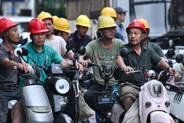 A group of construction workers on their scooters wait on a corner to cross a street after finishing their workday in Shanghai on August 13, 2026. (Photo by Hector RETAMAL / AFP)