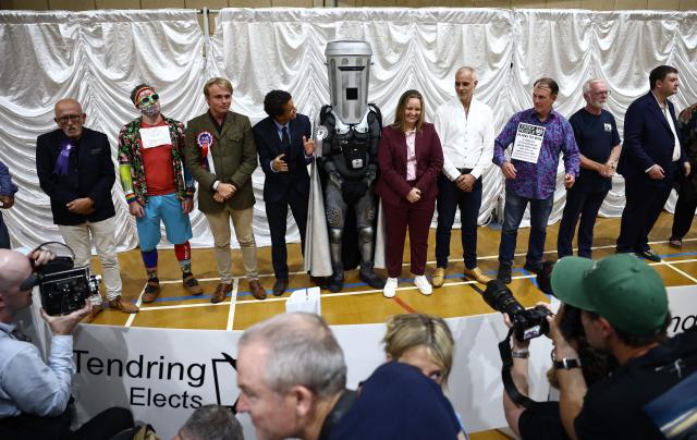 Candidates, including Count Binface (C), listen to the results for the by-election result for the parliamentary seat of Clacton, at the count centre venue, the Clacton Leisure Centre, eastern England on August 14, 2026. Hard-right British politician Nigel Farage of the Reform UK party, was declared on August 14, in an unusual by-election that pitted him against joke candidate Count Binface. (Photo by Henry NICHOLLS / AFP)