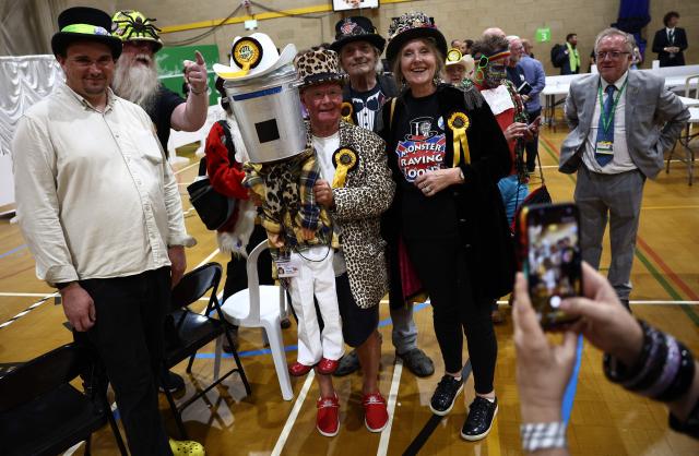 Members of the Monster Raving Looney Party bose with a character wearing a bin on its head, following the declaration of the Clacton by-election at Clacton Leisure Centre, eastern England on August 14, 2026. Hard-right British politician Nigel Farage of the Reform UK party, was declared on August 14, the winner of an unusual by-election that pitted him against joke candidate Count Binface. (Photo by Henry NICHOLLS / AFP)