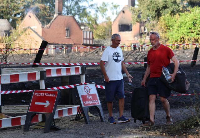 A person carries belongings in a suitcase, and a dog bed under their arm, as they walk near fire-damaged houses after a series of wildfires, in Stourbridge, central England on August 14, 2026. Firefighters continued Friday to extinguish wildfires that began on August 13, tearing through several homes, with residents evacuated and "extensive" damage reported. (Photo by Darren Staples / AFP)