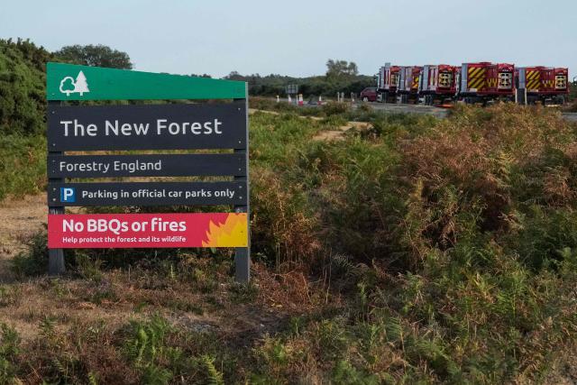 Fire engines (back R) are pictured behind a sign for the New Forest notifying visitors not to have BBQs or light fires, as wildfires have burned in the area for nearly five days, at Picket Post in southern England on August 14, 2026. Describing Britain as "a tinderbox" with 37 blazes nationwide on August 14, Britain's Prime Minister Andy Burnham warned against lighting fires outdoors, as the government introduced a ban on selling disposable barbecue equipment. (Photo by CARLOS JASSO / AFP)