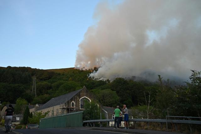 Local residents chat on the street as smoke rises from burning moorland near a residential area, south of Ebbw Vale in south Wales on August 15, 2026. Around 200 military personnel have been deployed to a large area across South Wales to help battle wildfires. (Photo by JUSTIN TALLIS / AFP)