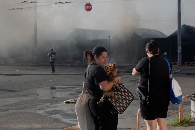 A woman holding a dog in her arms looks on as smoke rises in the background at the site of a Russian strike in Kyiv on August 16, 2026, amid the Russian invasion of Ukraine. Russian strikes killed one person and wounded at least seven others in Ukraine, authorities said August 16, while Moscow officials reported downing more than 100 drones heading towards the capital. (Photo by Tetiana DZHAFAROVA / AFP)