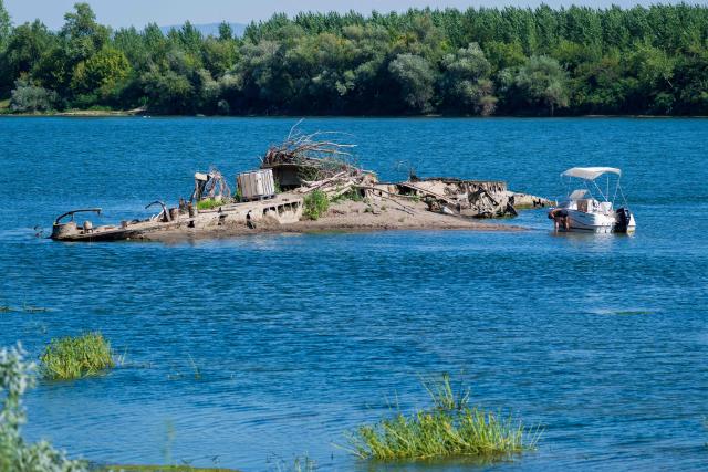 People visit the wreckage of the Slovenac, a paddle-wheel tugboat built in 1913, emerging from the low waters of the Sava River near Sremska Mitrovica, Serbia, on August 16, 2026, as drought and prolonged hot and dry weather continue to lower water levels across the region. According to Serbian media and local historians, the Slovenac was a paddle-wheel steam tug powered by two steam engines. It sank on April 25, 1945, after striking a German mine while transporting supplies and wounded soldiers during the final days of World War II, and has since become a local landmark that reappears during periods of exceptionally low water. (Photo by Andrej ISAKOVIC / AFP)