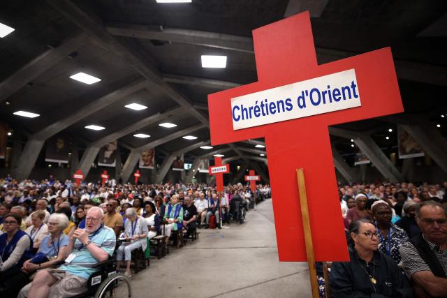 This photograph shows a faithful holding a cross bearing the message "Christians of the East" during Mass in the underground Basilica of St. Pius X at the Lourdes sanctuary, southwestern France, on August 16, 2026. (Photo by Valentine CHAPUIS / AFP)