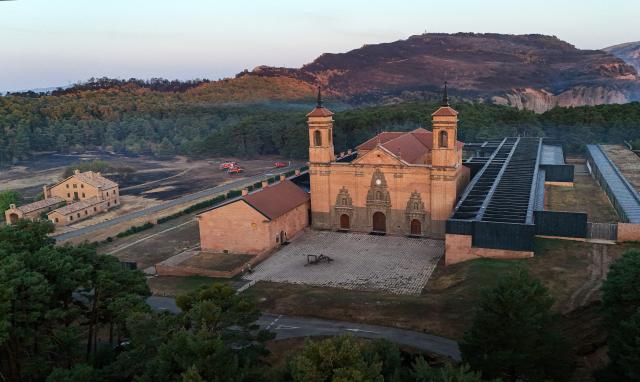 An aerial picture taken early on August 17, 2026 shows a view of the San Juan de la Pena Monastery in Penas de Riglos and burnt land as firefighters (L) are on guard to protect it from a wildfire, in the Spanish northeastern province of Aragon amid a wildfire that burnt 16,600 hectares. A Spanish soldier died on August 16, 2026 while helping efforts to extinguish a major wildfire in the northeastern region of Aragon. The fire that has raged for a week threatened a 1,000-year-old monastery, sheltering the bones of the first Aragonese kings, founders of the Kingdom of Aragon. (Photo by Oscar DEL POZO / AFP)
