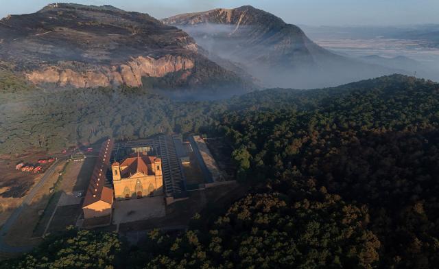 An aerial picture taken early on August 17, 2026 shows a view of the San Juan de la Pena Monastery and burnt land as firefighters (L) are on guard to protect it from a wildfire, in Penas de Riglos in the Spanish northeastern province of Aragon amid a wildfire that burnt 16,600 hectares. A Spanish soldier died on August 16, 2026 while helping efforts to extinguish a major wildfire in the northeastern region of Aragon. The fire that has raged for a week threatened a 1,000-year-old monastery, sheltering the bones of the first Aragonese kings, founders of the Kingdom of Aragon. (Photo by Oscar DEL POZO / AFP)