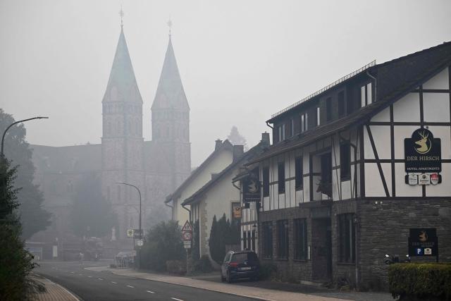 Smoke hangs on August 17, 2026 over the church and houses in  Kalterherberg near Monschau, western Germany, close to the border with Belgium. According to official reports, the fire in the Hohes Venn (High Fens, Hautes Fagnes) nature reserve in Belgium is no longer spreading towards the German border. "The wind is blowing in a favourable direction and the current rainfall is helping to contain the spread," said Tony Hosmans, head of crisis communications for the Governor of the Province of Liège, on Monday, August 17, 2026. Much-needed rain brought some respite Monday to firefighters on their fourth day battling Belgium's worst wildfire in a century, which has scorched vast expanses of nature reserve on the German border. Europe has been swept by weeks of devastating wildfires fuelled by dry conditions and a historically hot summer that has already contributed to the deaths of thousands of people. (Photo by INA FASSBENDER / AFP)