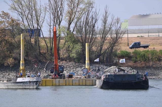 The controlled sinking of two 80-meter-long barges loaded with rocks is under way on the banks of the river Danube on the section of the Danube adjacent to the Paks nuclear power plant (background, R) near Dunaszentbenedek, Hungary, on August 18, 2026. Hungary has so far been able to avoid a complete shutdown of its only nuclear plant, Paks, whose four reactors are cooled by the Danube. But the Hungarian government has ordered the construction of a submerged wall, or weir, to try to control flows nears the plant. Two 80-metre barges are sunk to raise the water level. Paks, whose output usually covers around a third of Hungary's electricity needs, has been partially shut down since July 2026 due to a drought scorching Europe that has caused the Danube river to fall to record lows. (Photo by Attila KISBENEDEK / AFP)