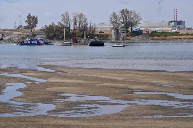 The controlled sinking of two 80-meter-long barges (C) loaded with rocks is under way on the banks of the river Danube on the section of the Danube adjacent to the Paks nuclear power plant (R) near Dunaszentbenedek, Hungary, on August 18, 2026. Hungary has so far been able to avoid a complete shutdown of its only nuclear plant, Paks, whose four reactors are cooled by the Danube. But the Hungarian government has ordered the construction of a submerged wall, or weir, to try to control flows nears the plant. Two 80-metre barges are sunk to raise the water level. Paks, whose output usually covers around a third of Hungary's electricity needs, has been partially shut down since July 2026 due to a drought scorching Europe that has caused the Danube river to fall to record lows. (Photo by Attila KISBENEDEK / AFP)
