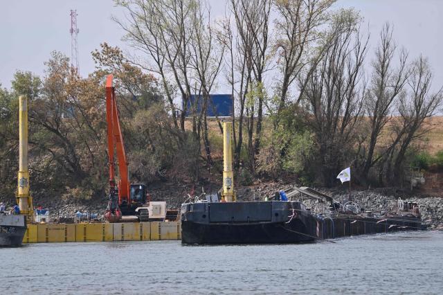 The controlled sinking of two 80-meter-long barges (R) loaded with rocks is under way on the banks of the river Danube on the section of the Danube adjacent to the Paks nuclear power plant near Dunaszentbenedek, Hungary, on August 18, 2026. Hungary has so far been able to avoid a complete shutdown of its only nuclear plant, Paks, whose four reactors are cooled by the Danube. But the Hungarian government has ordered the construction of a submerged wall, or weir, to try to control flows nears the plant. Two 80-metre barges are sunk to raise the water level. Paks, whose output usually covers around a third of Hungary's electricity needs, has been partially shut down since July 2026 due to a drought scorching Europe that has caused the Danube river to fall to record lows. (Photo by Attila KISBENEDEK / AFP)