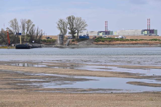 The controlled sinking of two 80-meter-long barges (L) loaded with rocks is under way on the banks of the river Danube on the section of the Danube adjacent to the Paks nuclear power plant (R) near Dunaszentbenedek, Hungary, on August 18, 2026. Hungary has so far been able to avoid a complete shutdown of its only nuclear plant, Paks, whose four reactors are cooled by the Danube. But the Hungarian government has ordered the construction of a submerged wall, or weir, to try to control flows nears the plant. Two 80-metre barges are sunk to raise the water level. Paks, whose output usually covers around a third of Hungary's electricity needs, has been partially shut down since July 2026 due to a drought scorching Europe that has caused the Danube river to fall to record lows. (Photo by Attila KISBENEDEK / AFP)