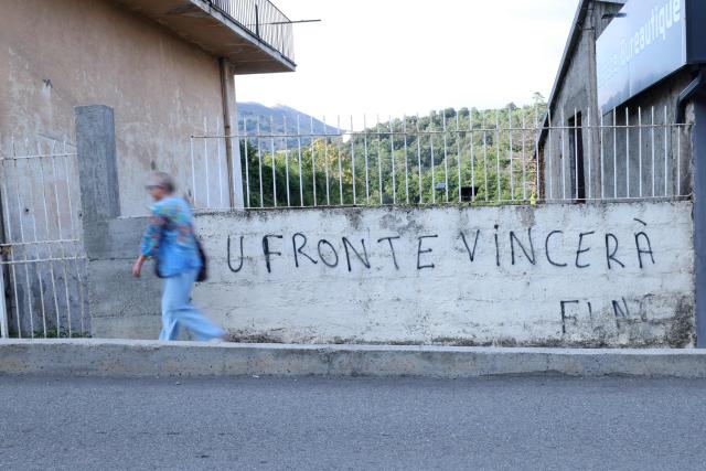 A woman walks past nationalist graffiti "The front will win - FLNC (Front de liberation nationale de la Corse)" on a wall in the town of Corte, in the center of the French Mediterranean island of Corsica, on August 19, 2026. (Photo by Pascal POCHARD-CASABIANCA / AFP)