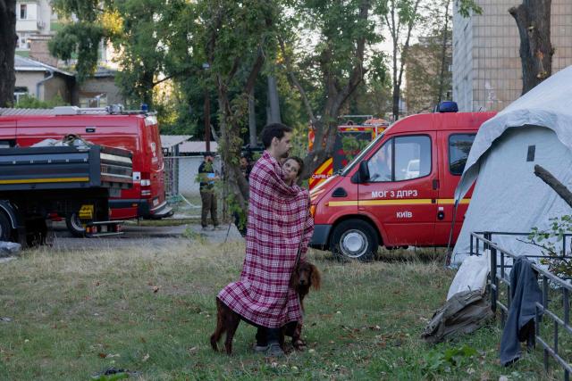 A man stands with a boy and a dog under a blanket near a damaged residential building following a Russian strike in Kyiv on August 20, 2026, amid Russia’s invasion of Ukraine. Russian bombardment killed six people and wounded dozens in Ukraine's capital and its surrounding region overnight, authorities said on August 20. (Photo by Tetiana DZHAFAROVA / AFP)