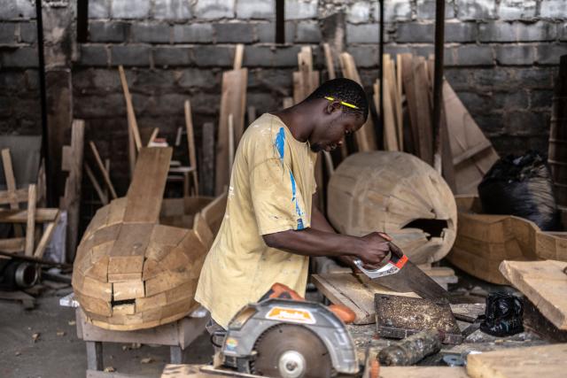 Coffin maker Lawrence Anang, 26, works on a shoe-shaped coffin, designed in reference to the profession of the deceased, a footballer, at the Kane Kwei Carpentry Workshop in Accra, on August 17, 2026. Fantasy coffins are a distinctive Ghanaian funerary tradition, particularly associated with the Ga people of the Greater Accra region. Known locally as abebuu adekai (“proverb boxes”), they are handcrafted in the shape of objects, animals or vehicles reflecting the deceased’s profession, personality or interests — from a fish for a fisherman to a shoe for a footballer. Developed around Accra in the mid-20th century, the tradition has evolved into a highly skilled craft. The practice reflects a Ghanaian approach to funerals in which mourning can coexist with celebration, symbolism and a strong emphasis on remembering the life and identity of the deceased. (Photo by OLYMPIA DE MAISMONT / AFP)