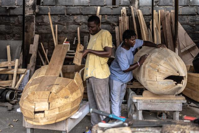 Coffin makers Lawrence Anang, 26, (L) and his brother Derrick Anang, 23, work on coffins at different stages of construction at the Kane Kwei Carpentry Workshop in Accra, on August 17, 2026. Fantasy coffins are a distinctive Ghanaian funerary tradition, particularly associated with the Ga people of the Greater Accra region. Known locally as abebuu adekai (“proverb boxes”), they are handcrafted in the shape of objects, animals or vehicles reflecting the deceased’s profession, personality or interests — from a fish for a fisherman to a shoe for a footballer. Developed around Accra in the mid-20th century, the tradition has evolved into a highly skilled craft. The practice reflects a Ghanaian approach to funerals in which mourning can coexist with celebration, symbolism and a strong emphasis on remembering the life and identity of the deceased. (Photo by OLYMPIA DE MAISMONT / AFP)