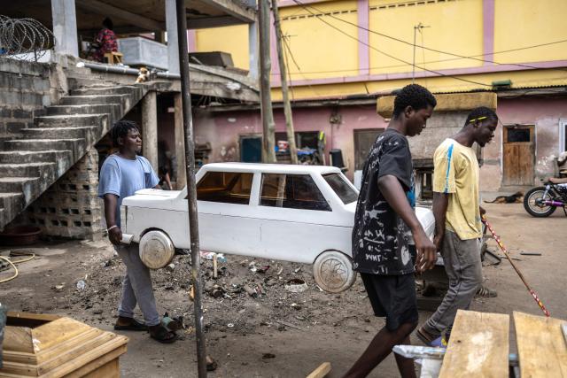 Coffin makers and brothers Lawrence Anang, 26, (R) Derrick Anang, 23, (L) and Ernest Anang, 18, (C) carry a car-shaped coffin, designed to replicate a car model owned by the deceased, at the Kane Kwei Carpentry Workshop in Accra, on August 17, 2026. Fantasy coffins are a distinctive Ghanaian funerary tradition, particularly associated with the Ga people of the Greater Accra region. Known locally as abebuu adekai (“proverb boxes”), they are handcrafted in the shape of objects, animals or vehicles reflecting the deceased’s profession, personality or interests — from a fish for a fisherman to a shoe for a footballer. Developed around Accra in the mid-20th century, the tradition has evolved into a highly skilled craft. The practice reflects a Ghanaian approach to funerals in which mourning can coexist with celebration, symbolism and a strong emphasis on remembering the life and identity of the deceased. (Photo by OLYMPIA DE MAISMONT / AFP)