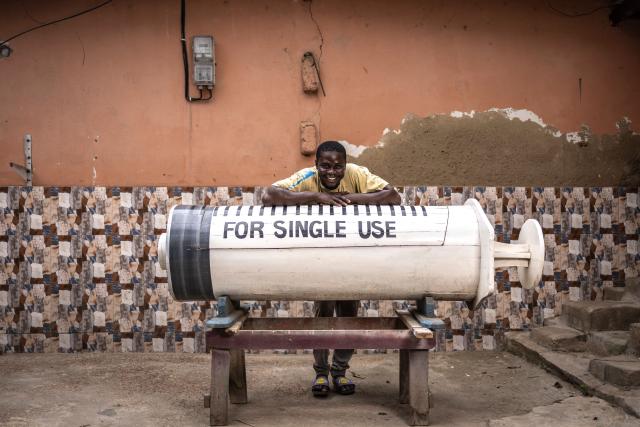 Coffin maker Lawrence Anang, 26, poses for a photograph with a syringe-shaped coffin, designed in reference to the profession of the deceased, a doctor, near the Kane Kwei Carpentry Workshop in Accra, on August 17, 2026. Fantasy coffins are a distinctive Ghanaian funerary tradition, particularly associated with the Ga people of the Greater Accra region. Known locally as abebuu adekai (“proverb boxes”), they are handcrafted in the shape of objects, animals or vehicles reflecting the deceased’s profession, personality or interests — from a fish for a fisherman to a shoe for a footballer. Developed around Accra in the mid-20th century, the tradition has evolved into a highly skilled craft. The practice reflects a Ghanaian approach to funerals in which mourning can coexist with celebration, symbolism and a strong emphasis on remembering the life and identity of the deceased. (Photo by OLYMPIA DE MAISMONT / AFP)