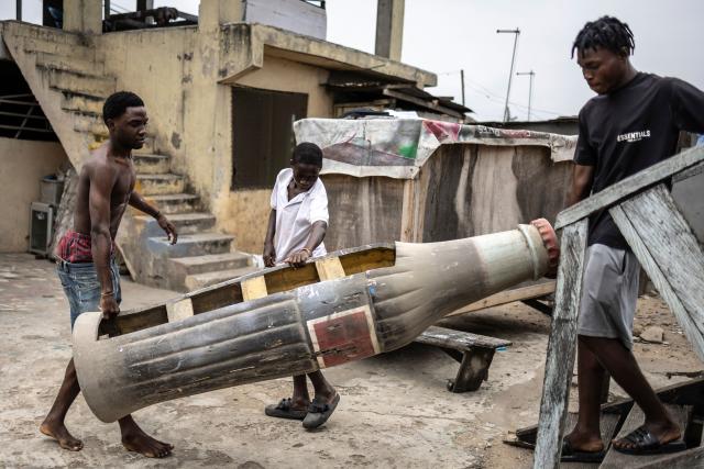 Men carry a Coca-Cola bottle-shaped coffin, designed in reference to the deceased's fondness for the soft drink, at the Hello Design Coffin workshop in Accra, on August 17, 2026. Fantasy coffins are a distinctive Ghanaian funerary tradition, particularly associated with the Ga people of the Greater Accra region. Known locally as abebuu adekai (“proverb boxes”), they are handcrafted in the shape of objects, animals or vehicles reflecting the deceased’s profession, personality or interests — from a fish for a fisherman to a shoe for a footballer. Developed around Accra in the mid-20th century, the tradition has evolved into a highly skilled craft. The practice reflects a Ghanaian approach to funerals in which mourning can coexist with celebration, symbolism and a strong emphasis on remembering the life and identity of the deceased. (Photo by OLYMPIA DE MAISMONT / AFP)