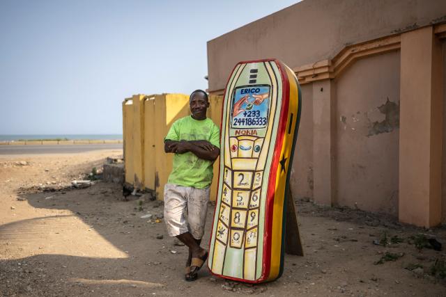 Eric Adotey Kpakpo, 49, also known as Erico, owner of the Erico Carpentry Shop, poses for a photograph next to a mobile phone-shaped coffin in Accra, on August 17, 2026. Fantasy coffins are a distinctive Ghanaian funerary tradition, particularly associated with the Ga people of the Greater Accra region. Known locally as abebuu adekai (“proverb boxes”), they are handcrafted in the shape of objects, animals or vehicles reflecting the deceased’s profession, personality or interests — from a fish for a fisherman to a shoe for a footballer. Developed around Accra in the mid-20th century, the tradition has evolved into a highly skilled craft. The practice reflects a Ghanaian approach to funerals in which mourning can coexist with celebration, symbolism and a strong emphasis on remembering the life and identity of the deceased. (Photo by OLYMPIA DE MAISMONT / AFP)