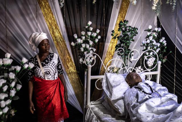 A mourner stands next to the body of late Ghanaian teacher Samuel Commey Tetteh during his funeral ceremony in Paanor, Ghana, on August 15, 2026. Fantasy coffins are a distinctive Ghanaian funerary tradition, particularly associated with the Ga people of the Greater Accra region. Known locally as abebuu adekai (“proverb boxes”), they are handcrafted in the shape of objects, animals or vehicles reflecting the deceased’s profession, personality or interests — from a fish for a fisherman to a shoe for a footballer. Developed around Accra in the mid-20th century, the tradition has evolved into a highly skilled craft. The practice reflects a Ghanaian approach to funerals in which mourning can coexist with celebration, symbolism and a strong emphasis on remembering the life and identity of the deceased. (Photo by OLYMPIA DE MAISMONT / AFP)
