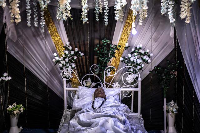 The body of the late Ghanaian teacher Samuel Commey Tetteh is displayed during his funeral ceremony in Paanor, Ghana, on August 15, 2026. Fantasy coffins are a distinctive Ghanaian funerary tradition, particularly associated with the Ga people of the Greater Accra region. Known locally as abebuu adekai (“proverb boxes”), they are handcrafted in the shape of objects, animals or vehicles reflecting the deceased’s profession, personality or interests — from a fish for a fisherman to a shoe for a footballer. Developed around Accra in the mid-20th century, the tradition has evolved into a highly skilled craft. The practice reflects a Ghanaian approach to funerals in which mourning can coexist with celebration, symbolism and a strong emphasis on remembering the life and identity of the deceased. (Photo by OLYMPIA DE MAISMONT / AFP)