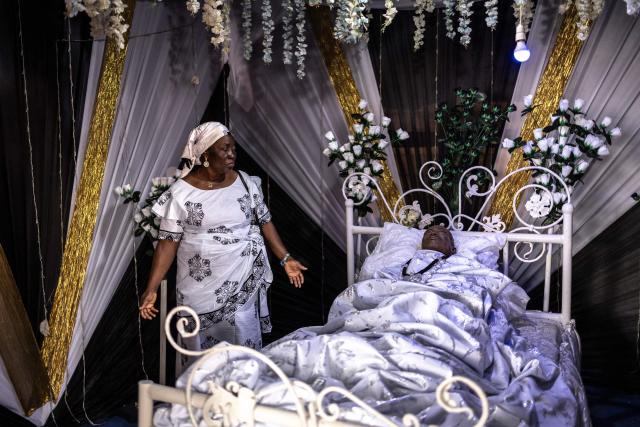 A mourner stands next to a body of the late Ghanaian teacher Samuel Commey Tetteh during his funeral ceremony in Paanor, Ghana, on August 15, 2026. Fantasy coffins are a distinctive Ghanaian funerary tradition, particularly associated with the Ga people of the Greater Accra region. Known locally as abebuu adekai (“proverb boxes”), they are handcrafted in the shape of objects, animals or vehicles reflecting the deceased’s profession, personality or interests — from a fish for a fisherman to a shoe for a footballer. Developed around Accra in the mid-20th century, the tradition has evolved into a highly skilled craft. The practice reflects a Ghanaian approach to funerals in which mourning can coexist with celebration, symbolism and a strong emphasis on remembering the life and identity of the deceased. (Photo by OLYMPIA DE MAISMONT / AFP)