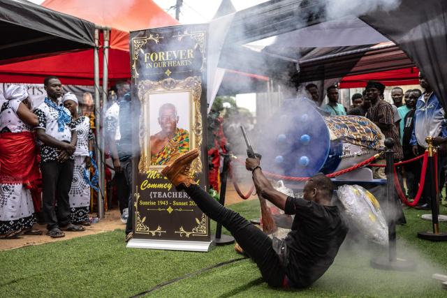 A man fires a traditional firearm next to the pen-shaped coffin of the late Ghanaian teacher Samuel Commey Tetteh during his funeral ceremony in Paanor, Ghana, on August 15, 2026. Fantasy coffins are a distinctive Ghanaian funerary tradition, particularly associated with the Ga people of the Greater Accra region. Known locally as abebuu adekai (“proverb boxes”), they are handcrafted in the shape of objects, animals or vehicles reflecting the deceased’s profession, personality or interests — from a fish for a fisherman to a shoe for a footballer. Developed around Accra in the mid-20th century, the tradition has evolved into a highly skilled craft. The practice reflects a Ghanaian approach to funerals in which mourning can coexist with celebration, symbolism and a strong emphasis on remembering the life and identity of the deceased. (Photo by OLYMPIA DE MAISMONT / AFP)