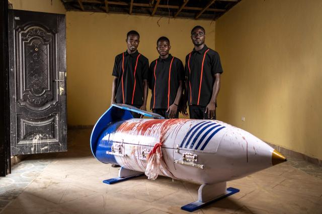 Relatives Eric Commey, Benjamin Commey and Ernest Commey pose for a photograph with the sheep blood-stained pen-shaped coffin of the late Ghanaian teacher Samuel Commey Tetteh inside his home in his native village of Akotuaku, on August 15, 2026. Fantasy coffins are a distinctive Ghanaian funerary tradition, particularly associated with the Ga people of the Greater Accra region. Known locally as abebuu adekai (“proverb boxes”), they are handcrafted in the shape of objects, animals or vehicles reflecting the deceased’s profession, personality or interests — from a fish for a fisherman to a shoe for a footballer. Developed around Accra in the mid-20th century, the tradition has evolved into a highly skilled craft. The practice reflects a Ghanaian approach to funerals in which mourning can coexist with celebration, symbolism and a strong emphasis on remembering the life and identity of the deceased. (Photo by OLYMPIA DE MAISMONT / AFP)